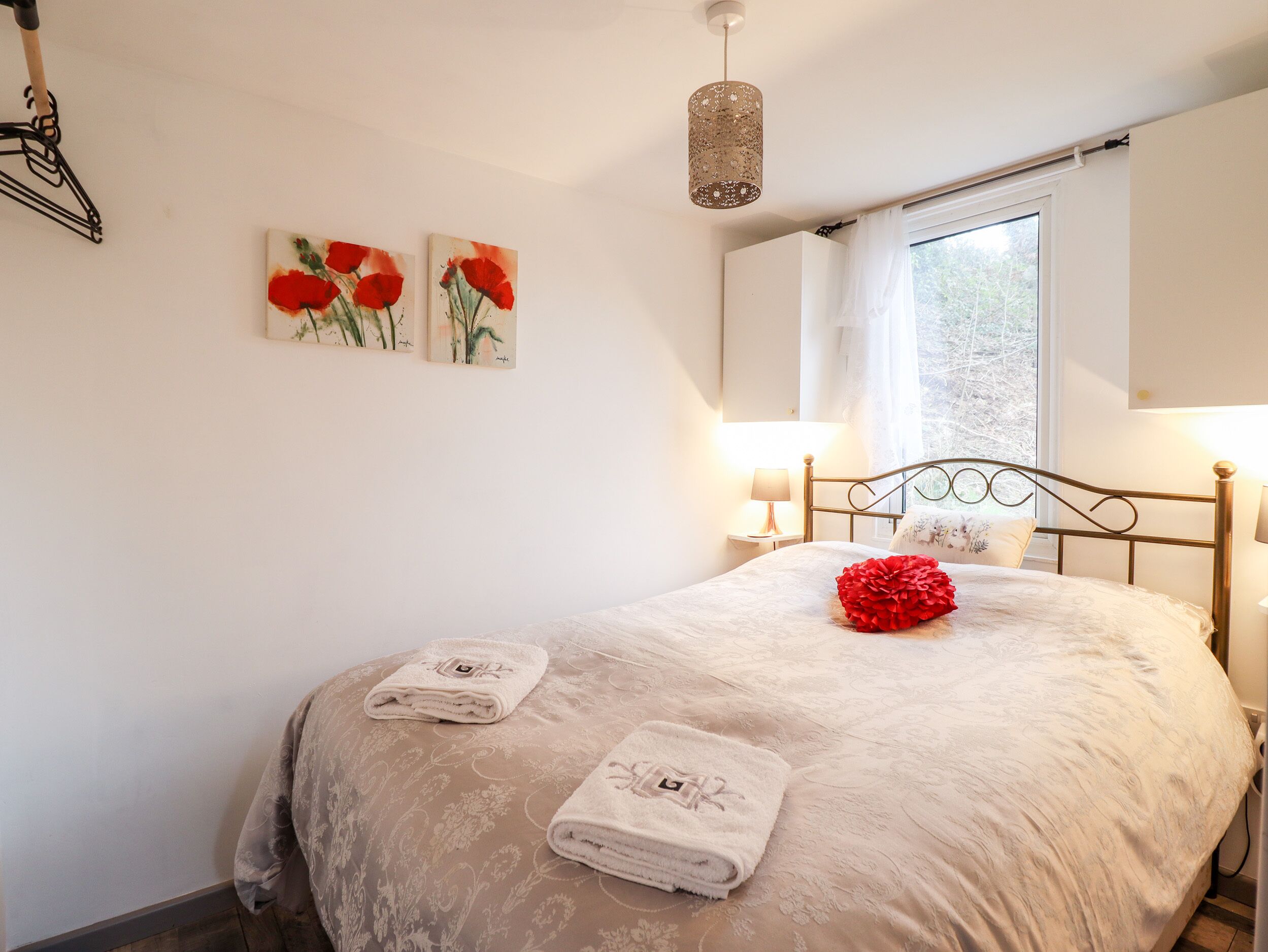 A bedroom with a bed, two wall-mounted lamps, a window with curtains, and two paintings of red flowers.
