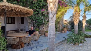 A man sitting at a wooden table with wicker chairs under a thatched roof, surrounded by lush greenery and palm trees. - Divers House (Mykonos)