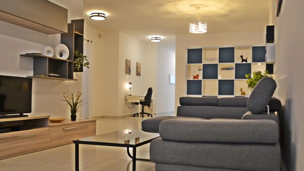 Ein modernes Wohnzimmer mit grauem Sofa, einem Glastisch und einem Fernseher auf einer Holzkommode. Regale mit Dekorationselementen und ein Schreibtisch mit Stuhl sind ebenfalls vorhanden.