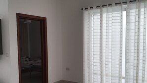 Ein Zimmer mit Holztür, Bett, weißen Wänden und einem Fenster mit durchsichtigen Vorhängen.