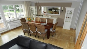Uma sala de jantar com mesa de madeira e cadeiras de vime, um sofá de couro preto e vista para um jardim através de grandes janelas.