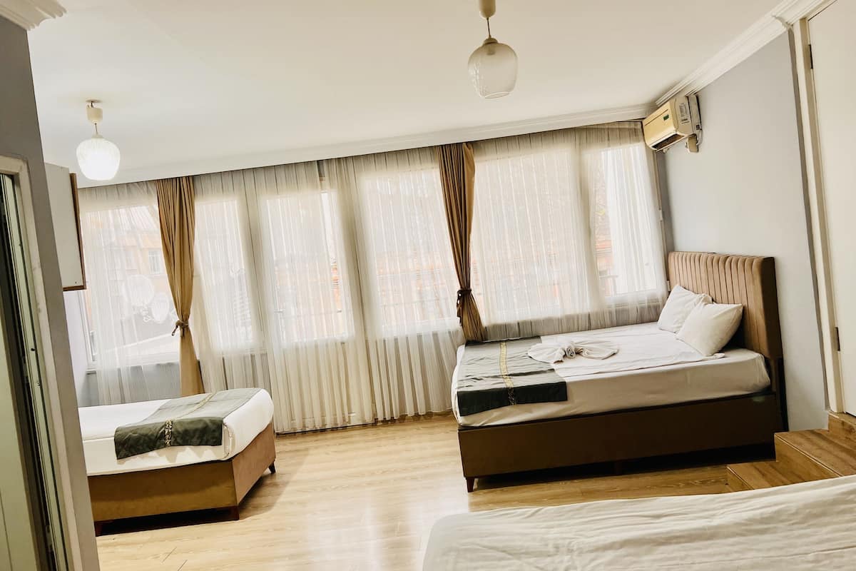 A hotel room with two beds, a large window with sheer curtains, a ceiling light, and a wall-mounted air conditioning unit.