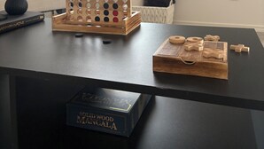 Ett vardagsrum med ett soffbord, ett träspelbräde och en låda märkt "Mancala i massivt trä". En växt och en soffa syns i bakgrunden.