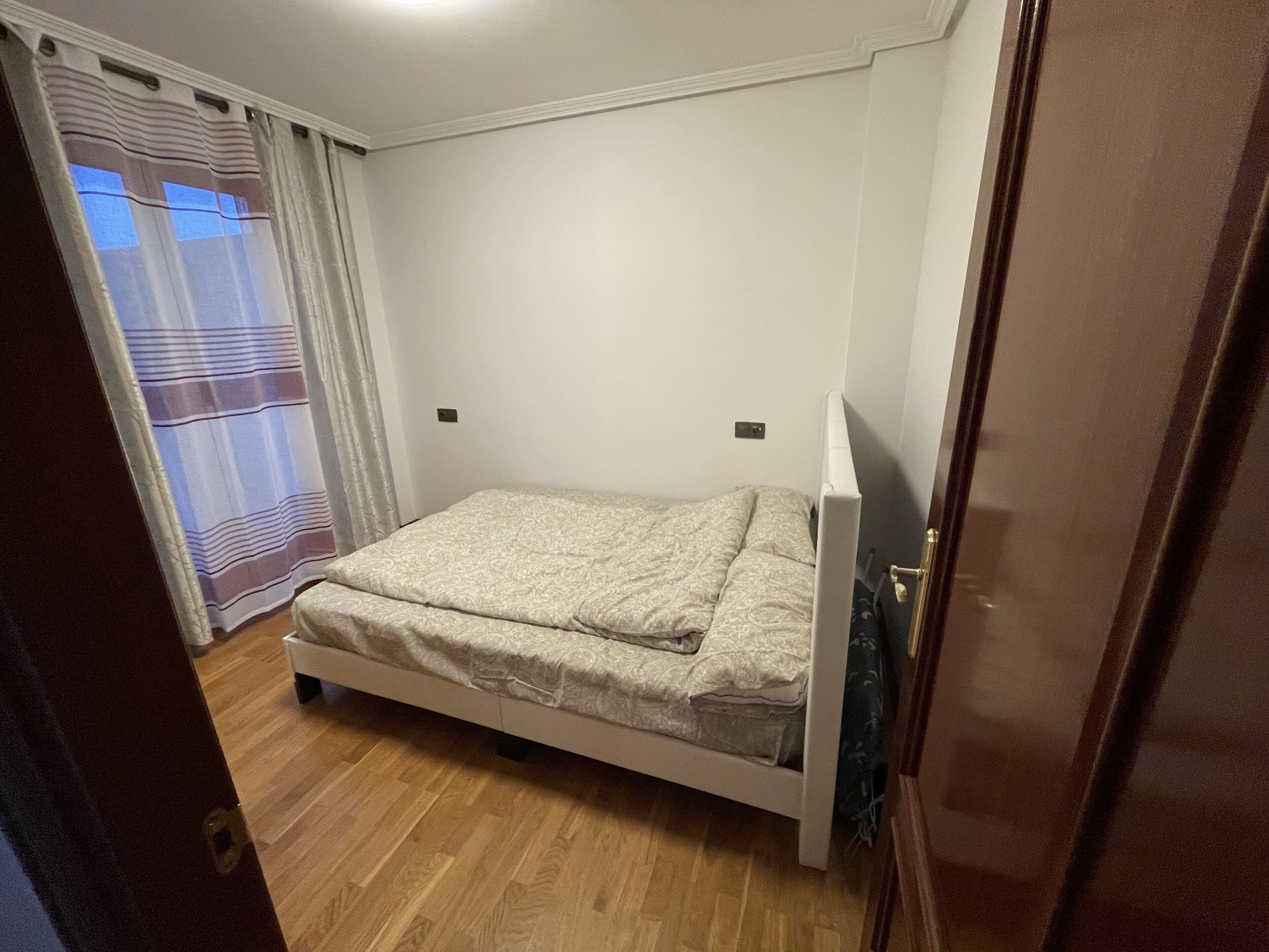 A bedroom with a bed, wooden flooring, a window with curtains, and a door.