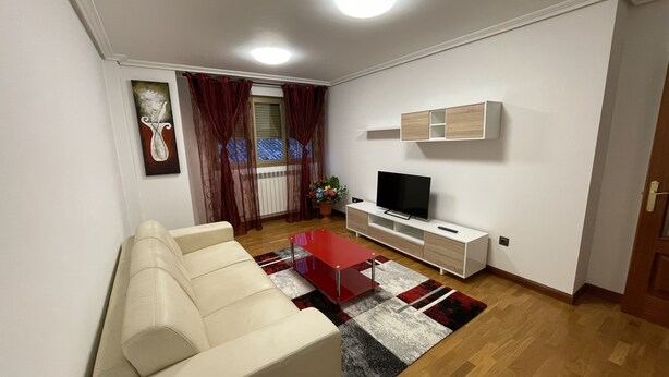 A living room with a sofa, a TV on a white stand, a red coffee table, and a patterned rug.