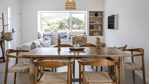 A dining area with a wooden table and chairs, a woven pendant light, and a view of a balcony with a sofa and cushions. - Casa do Cacto - By Wehost (Porto Santo)