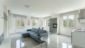 A modern living room with a grey sectional sofa, a small white side table, and a kitchen area with a built-in oven and a sink.