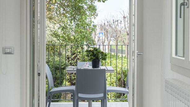 An open door to a balcony with a table and chairs, a potted plant, and a view of trees and buildings.
