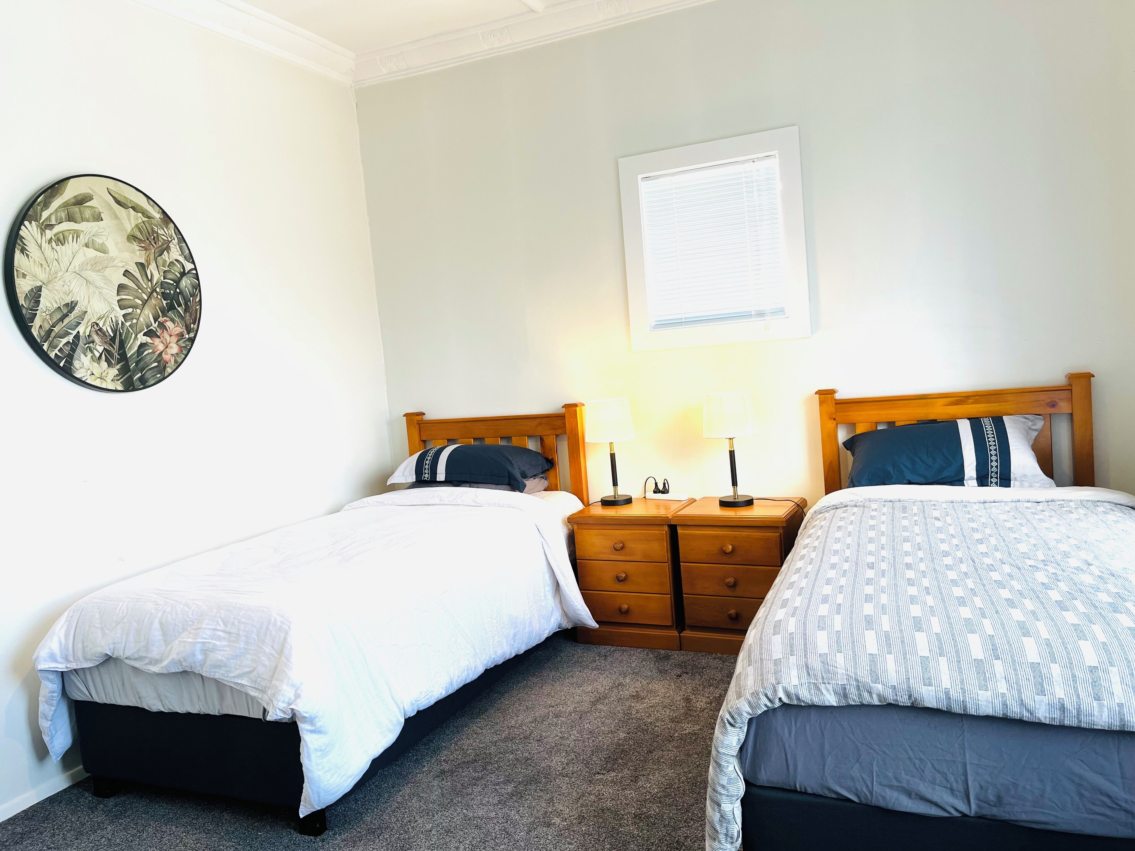 A bedroom with two single beds, a nightstand with a lamp, a wall-mounted picture, and a window with blinds.