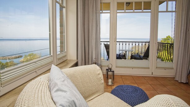 A room with a large window overlooking a body of water, a balcony with outdoor seating, and a cozy seating area with cushions. - Vacation apartment with fantastic sea view - Haus am Meer FeWo 05 (Lohme)