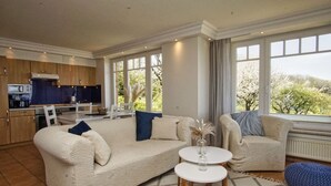 A spacious living area with a large beige sofa, a smaller sofa, and a round coffee table. There is a kitchen area with wooden cabinets and a dining table. Large windows provide a view of trees and a building. - Vacation apartment with fantastic sea view - Haus am Meer FeWo 05 (Lohme)