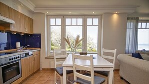A kitchen with wooden cabinets, a stove, and a dining table with chairs. There is a window with a view of trees and a body of water. - Vacation apartment with fantastic sea view - Haus am Meer FeWo 05 (Lohme)