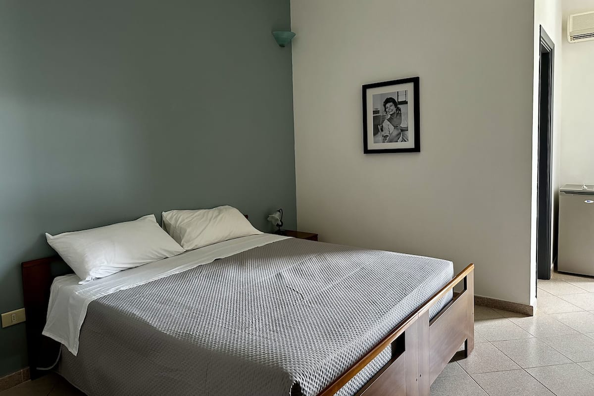Een slaapkamer met een bed, een nachtkastje, een lijst met een afbeelding aan de muur en een deur naar een andere kamer.