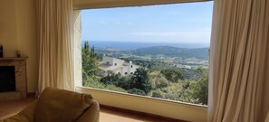 A room with a large window offering a view of a coastal landscape, a fireplace, and a beige sofa.