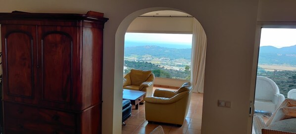 A room with a view of a valley and mountains, a wooden cabinet, and two chairs.
