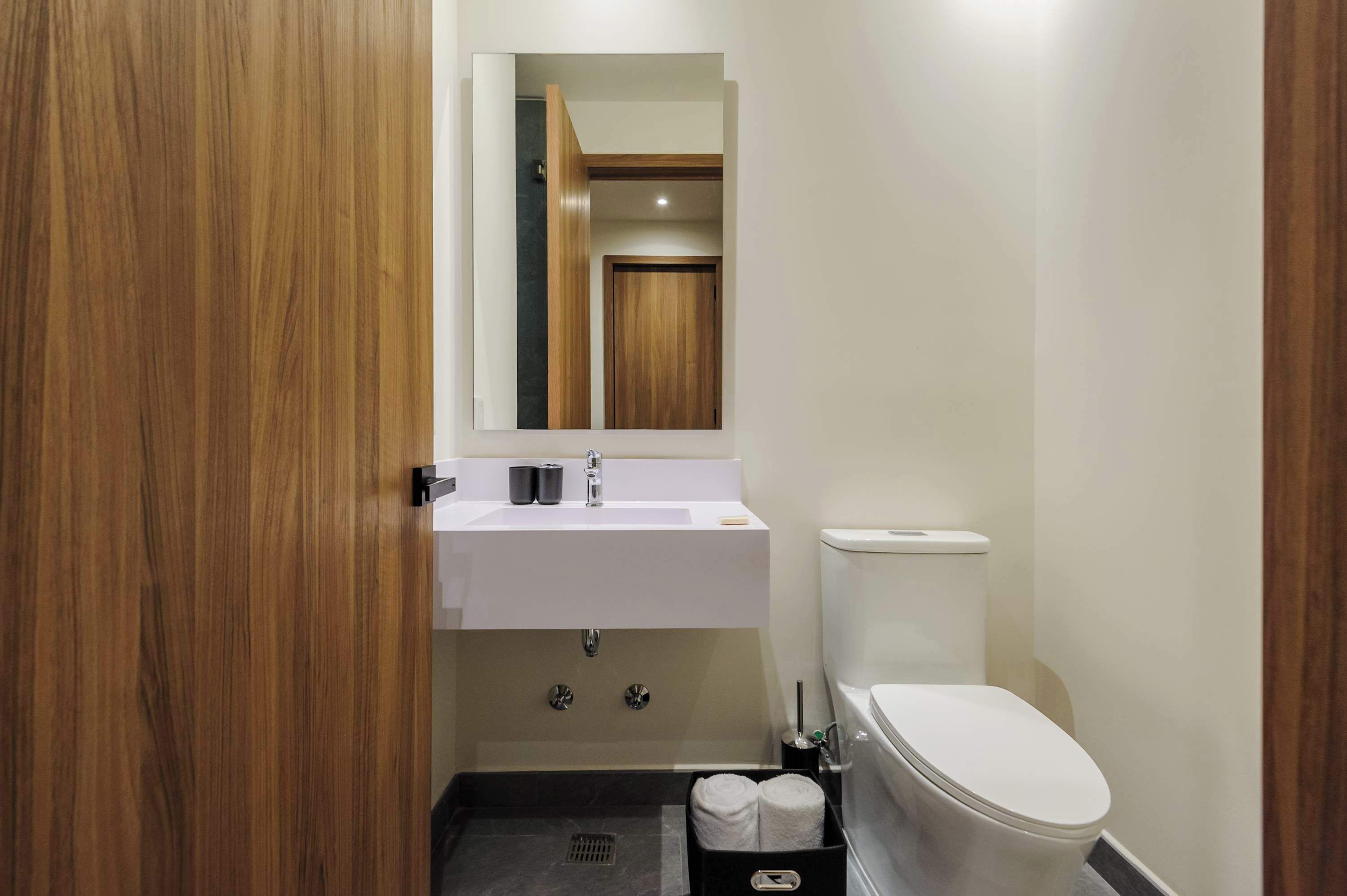 Une salle de bain moderne avec une porte en bois, un lavabo blanc, un miroir, des toilettes et une poubelle noire.