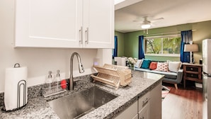 A kitchen with a granite countertop, stainless steel sink, and white cabinets. There is a wooden rack for utensils, a paper towel holder, and a ceiling fan. - Uhost AI | Hollywood Garden 1BR Family Spot (Hollywood)