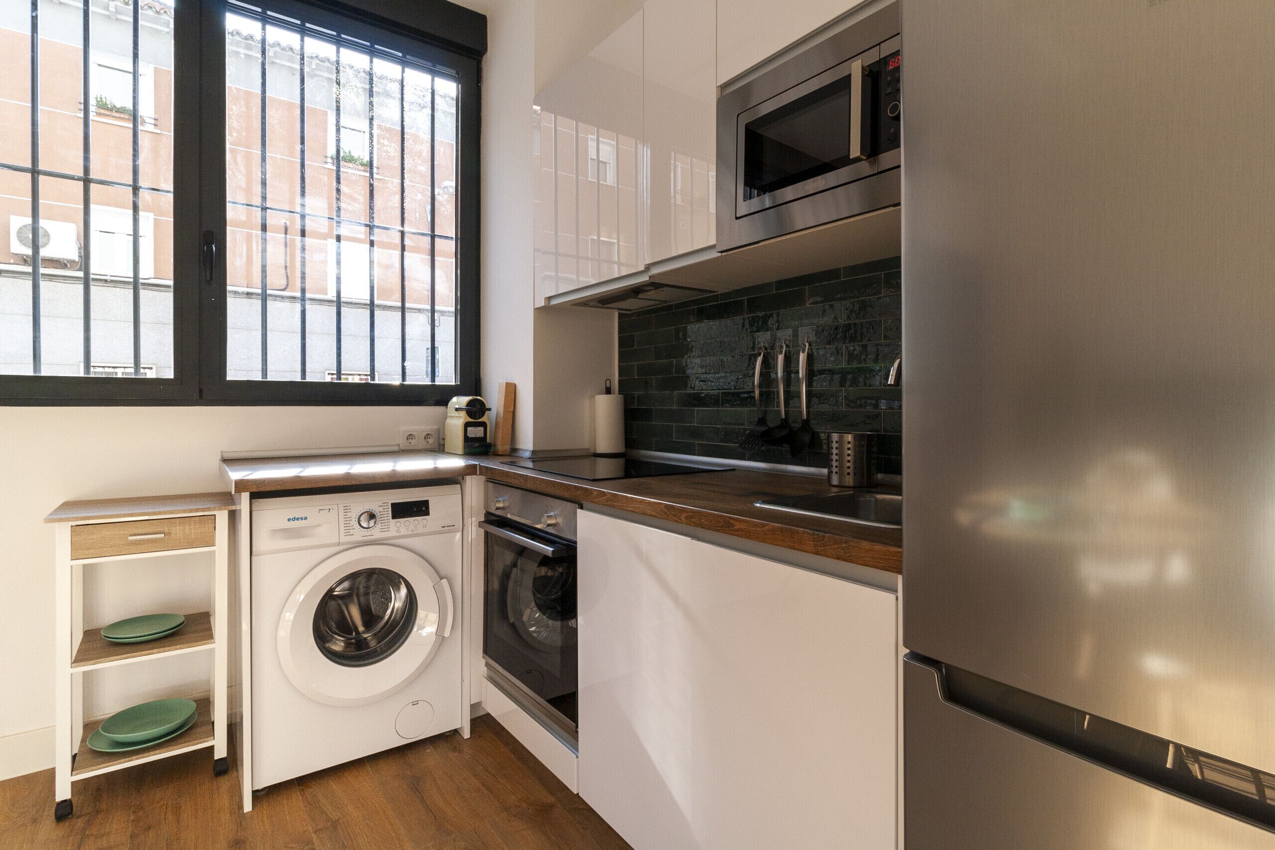A compact kitchen with a stainless steel refrigerator, a built-in oven, a washing machine, and a microwave.