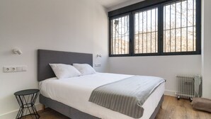 A modern bedroom with a large bed, a small side table, a radiator, and a window with bars.