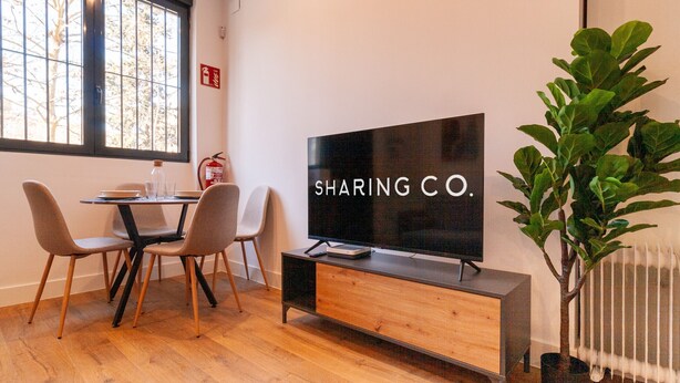 A modern living room with a large flat-screen TV, a dining table with chairs, and a potted plant.