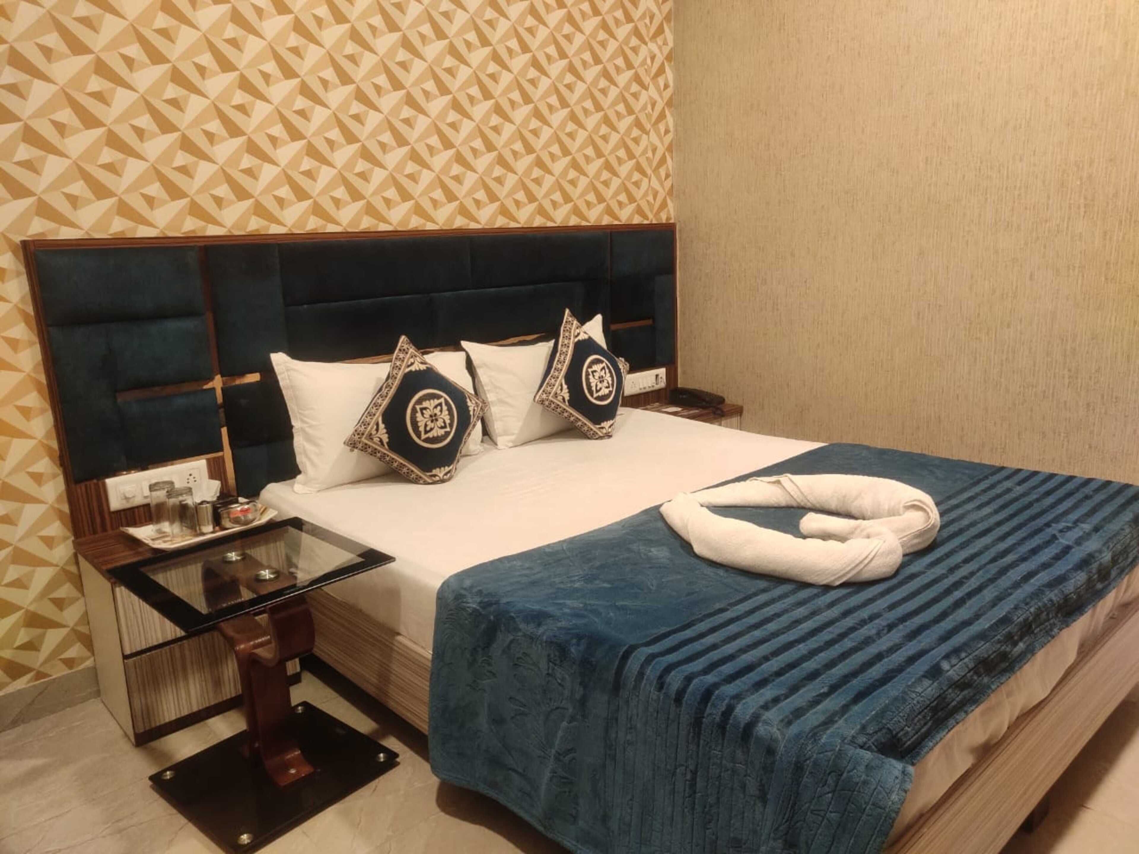 A neatly made bed with a blue blanket and decorative pillows, a glass side table with a tray of items, and a patterned wall in the background.