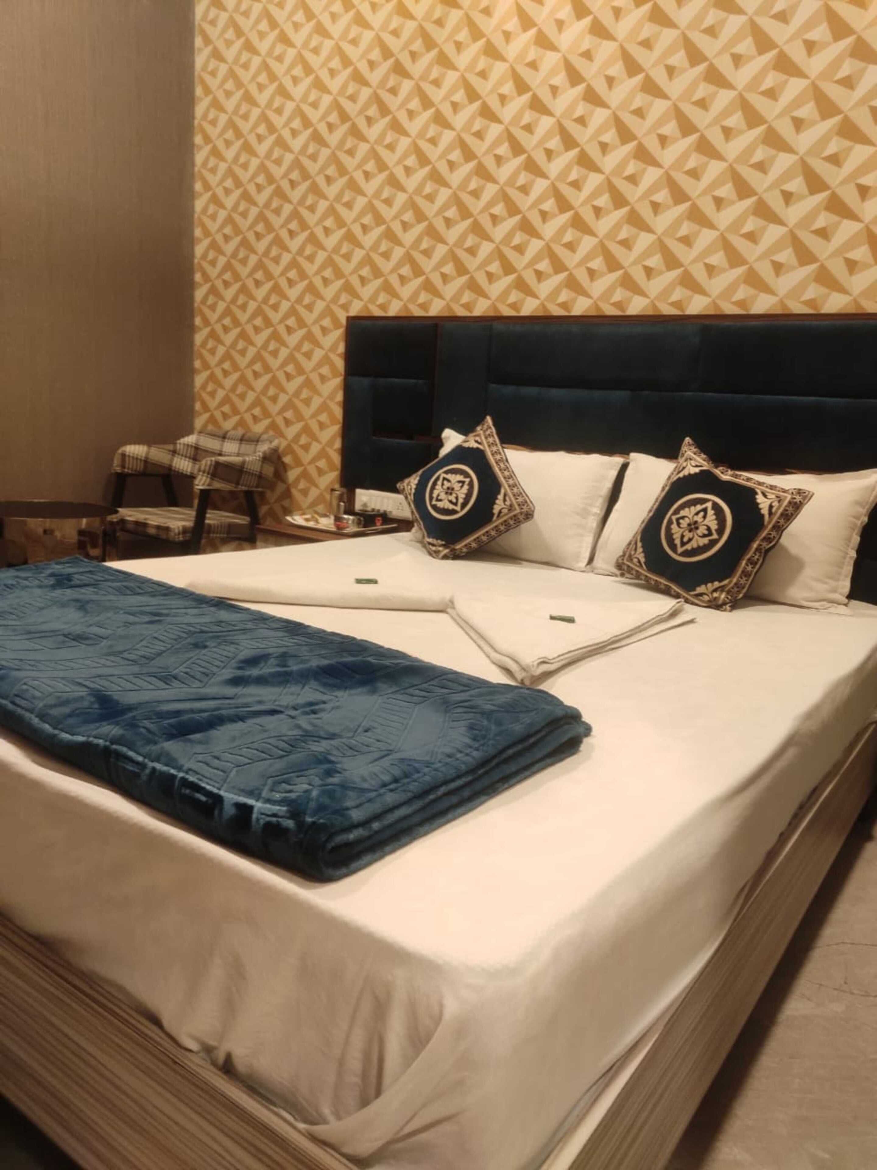 Una cama bien hecha con una manta azul y dos almohadas decorativas, un sillón escocés y un papel tapiz estampado en una habitación de hotel.