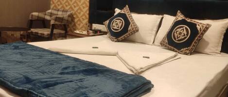 A neatly made bed with a blue blanket and two decorative pillows, a plaid chair, and a patterned wall in a hotel room.