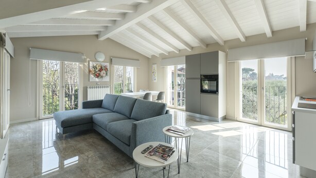 Ein geräumiges Wohnzimmer mit einem grauen Ecksofa, einem kleinen Couchtisch und einem an der Wand befestigten Fernseher. Große Fenster sorgen für viel natürliches Licht und Ausblick ins Freie.