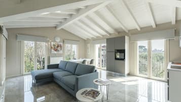 Un soggiorno spazioso con un divano modulare grigio, un tavolino da caffè e una TV fissata al muro. Grandi finestre offrono molta luce naturale e vista sull'esterno.