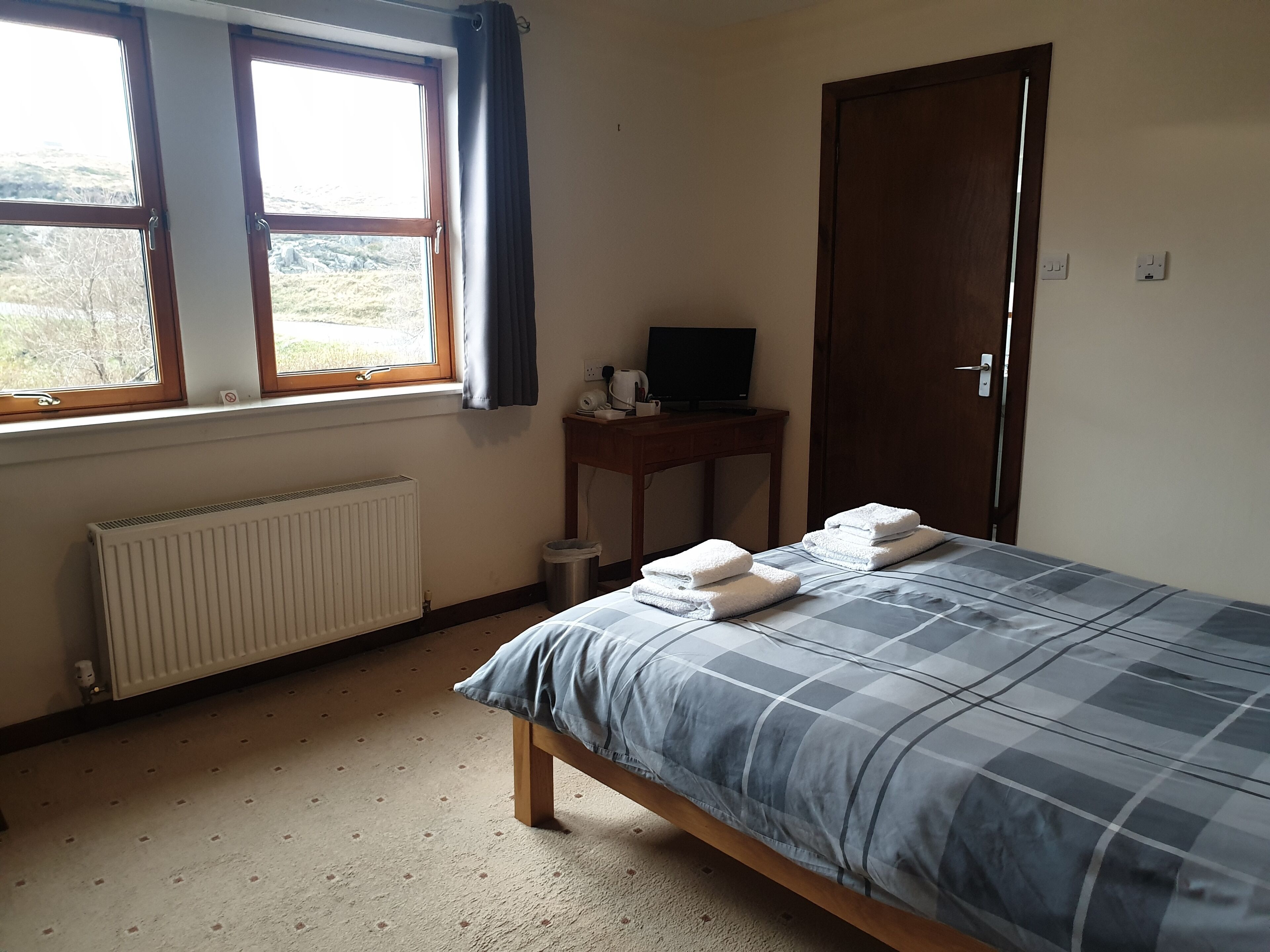 Une chambre à coucher avec un lit, un radiateur, une fenêtre avec des rideaux, un bureau avec une télévision et une porte.