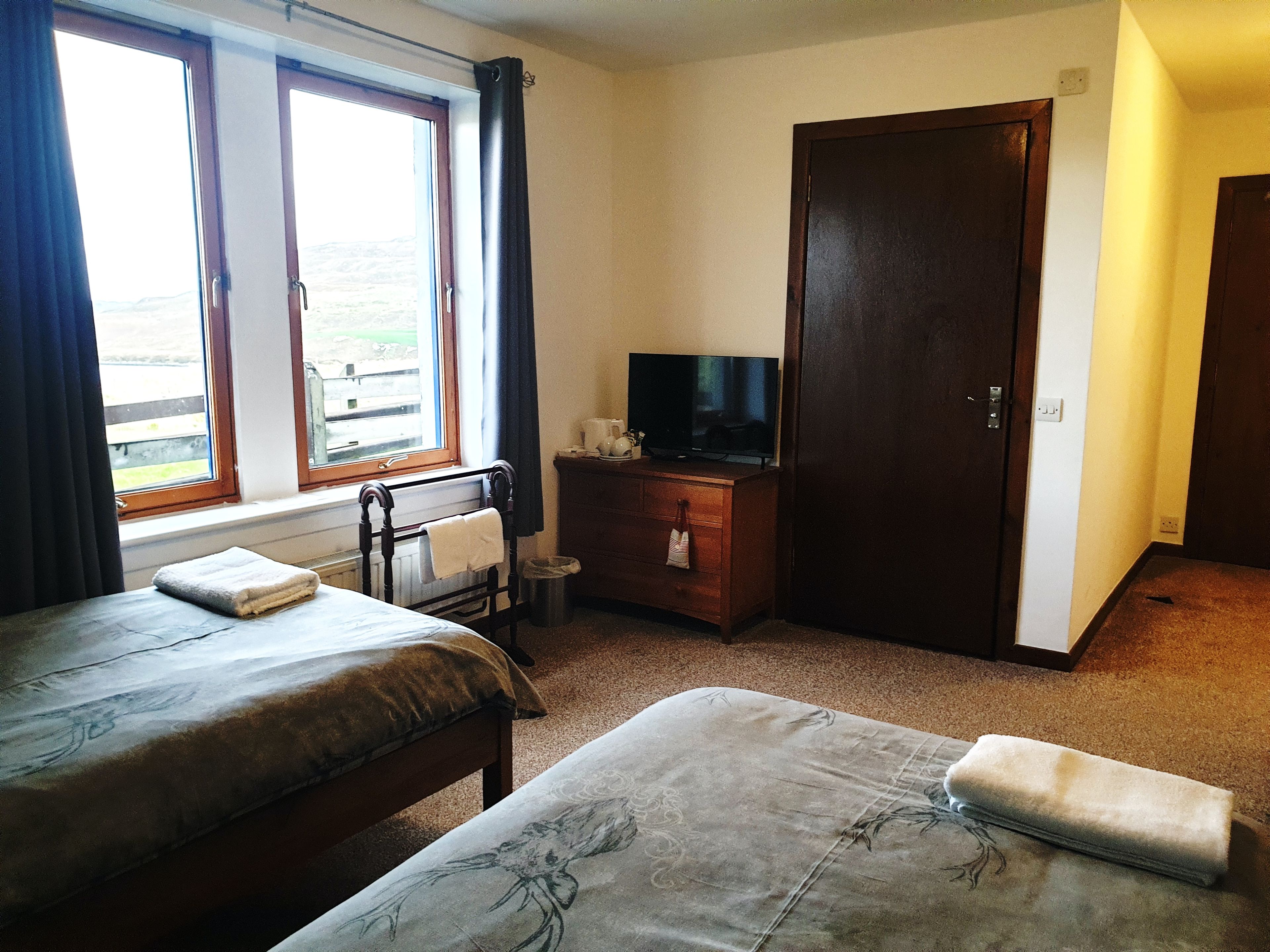 A room with two beds, a television on a dresser, a window with curtains, and a door.