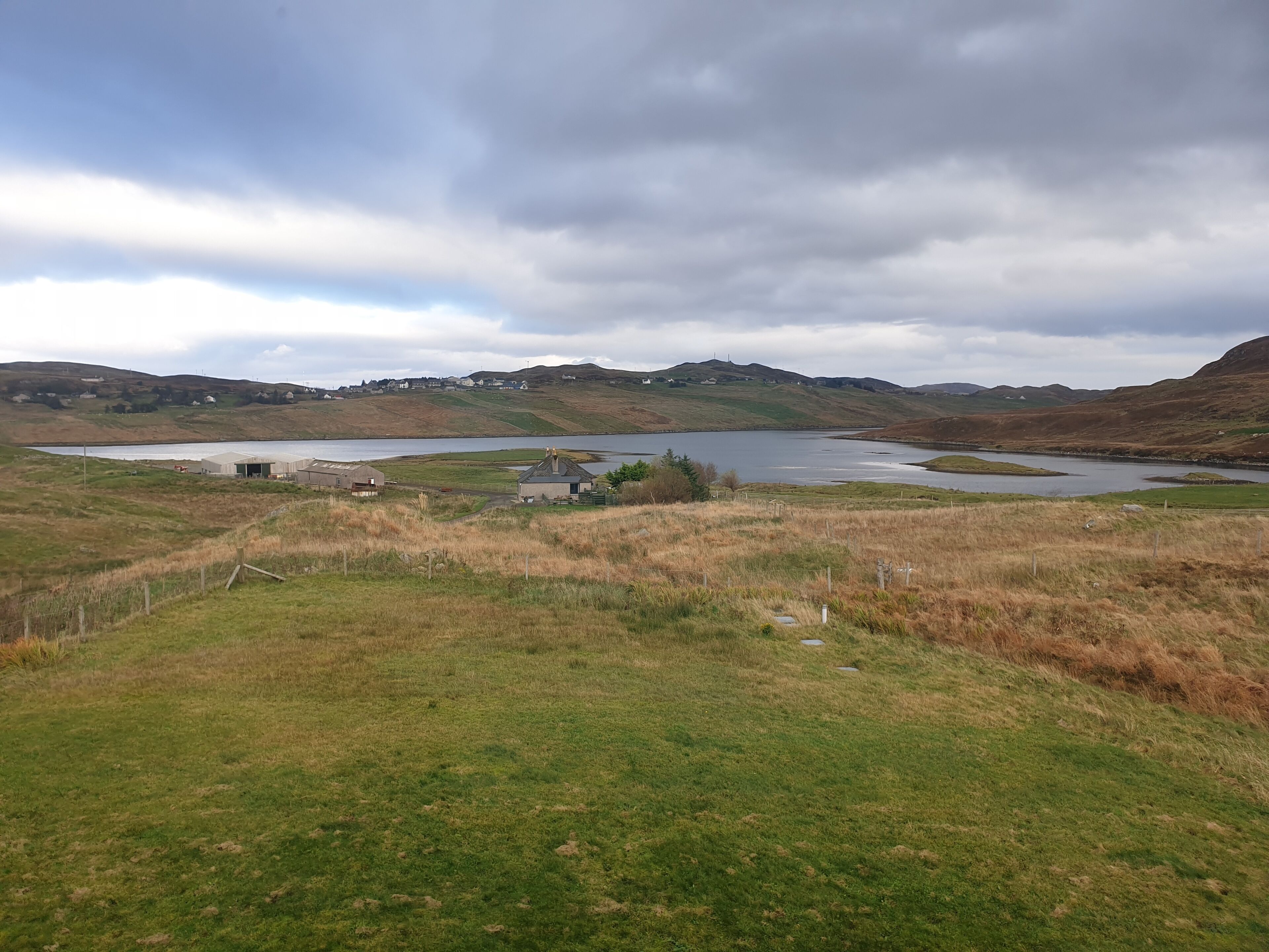 Un paysage rural avec un lac, des champs verdoyants et quelques bâtiments visibles au loin.