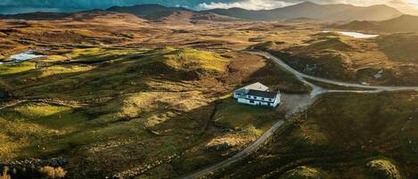 A remote house with a dirt road leading to it, surrounded by rolling hills and a lake.