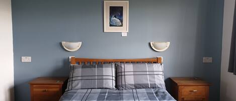 Uma cama bem arrumada com colcha xadrez, duas mesas de cabeceira e um quadro na parede.