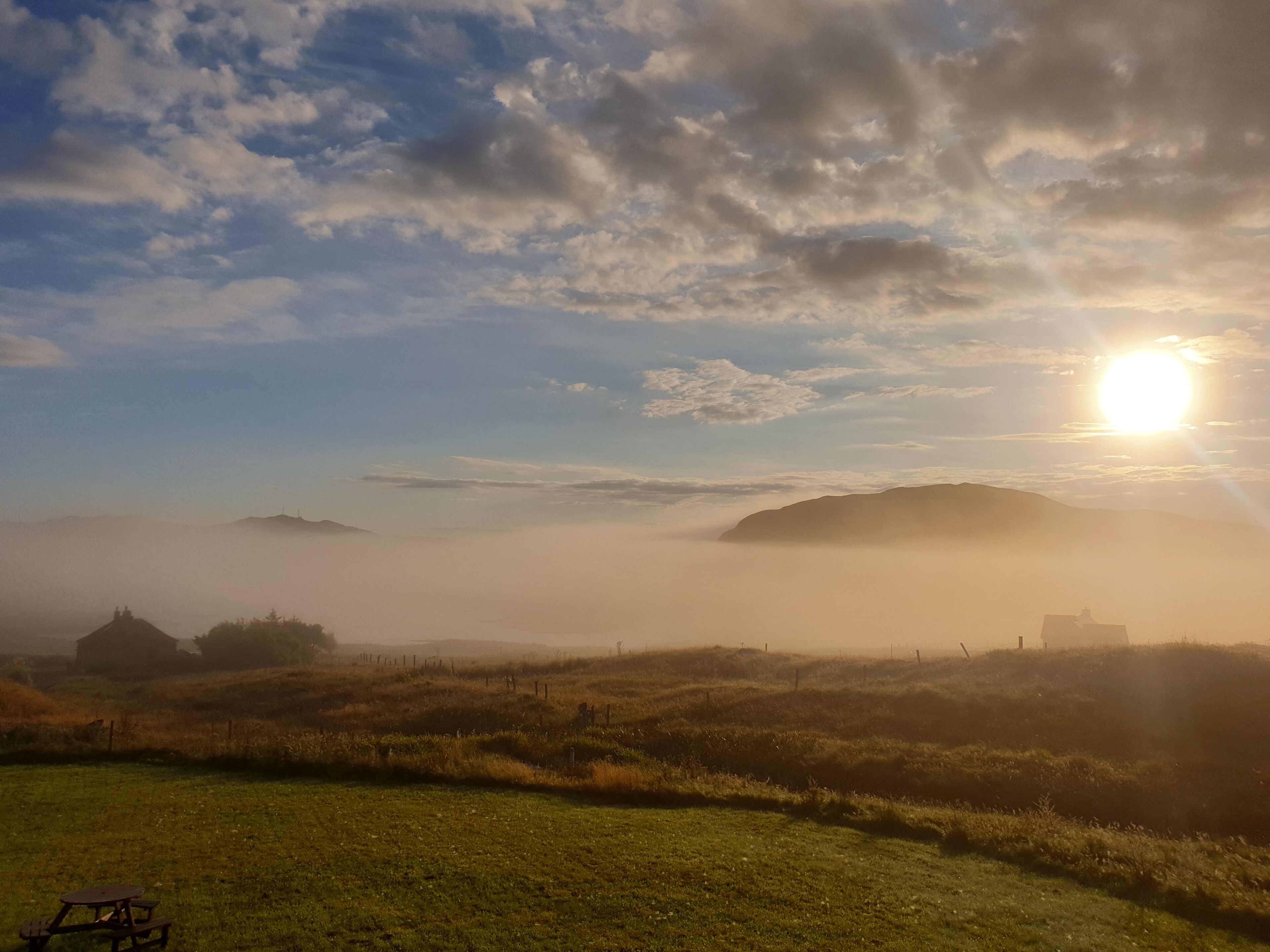 Un paysage brumeux, avec un soleil couchant derrière des collines, une table de pique-nique et un bâtiment situé au loin.