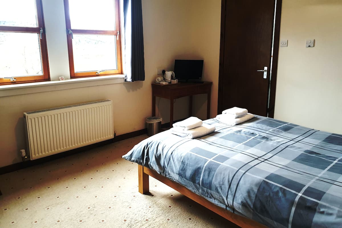 A bedroom with a bed, a radiator, a window with curtains, a desk with a television, and a door.