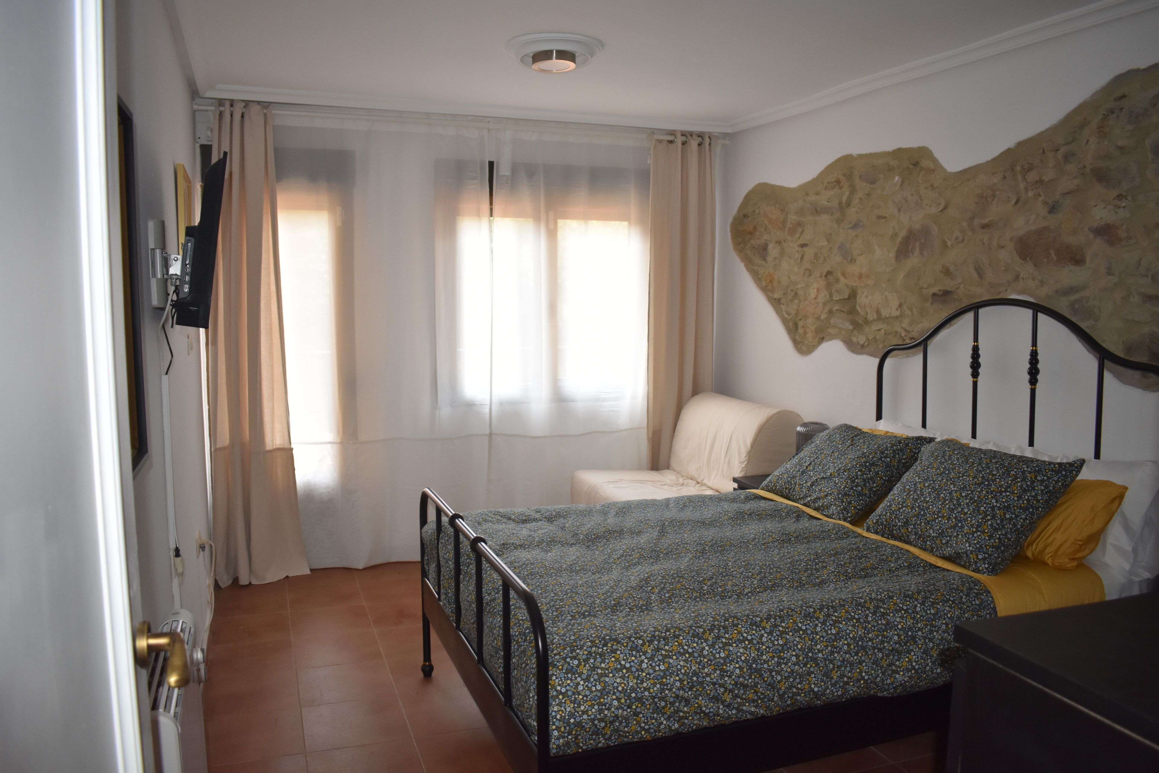 Une chambre à coucher avec un lit, une chaise, un mur en pierre et une fenêtre avec des rideaux.