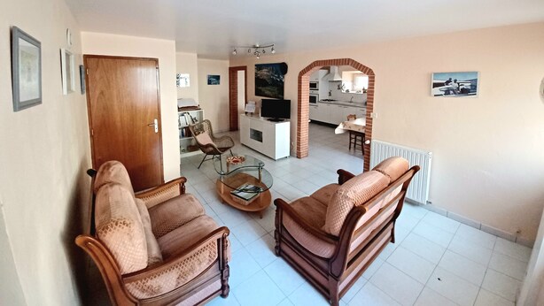A living room with two brown wicker sofas, a coffee table, and a TV. A kitchen is visible through an archway in the background.