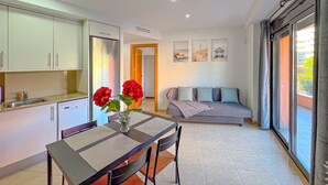 A modern kitchen with white cabinets, a dining table with chairs, a vase with red flowers, and a living area with a sofa and a window with a view. - Apartment for 3 people, free Wi-Fi in the center of Lloret, 300 meters from the beach, ideal for young people. (Lloret de Mar)