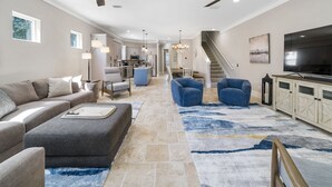 Ein geräumiges Wohnzimmer mit einer Couch, Sesseln, einem Couchtisch und einem Fernseher. Im Hintergrund befindet sich eine Küche, eine Treppe und ein Chandelier.