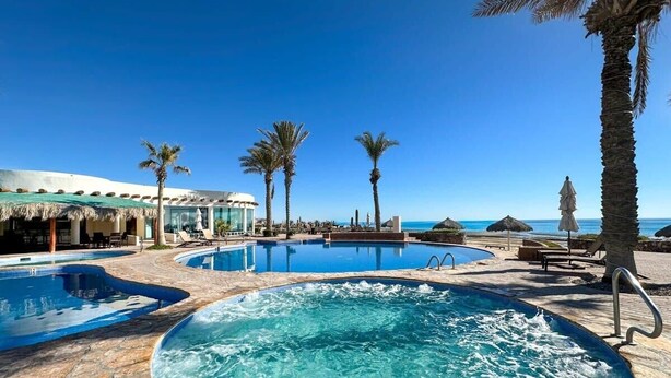 A swimming pool area with multiple pools, palm trees, and a beach view.
