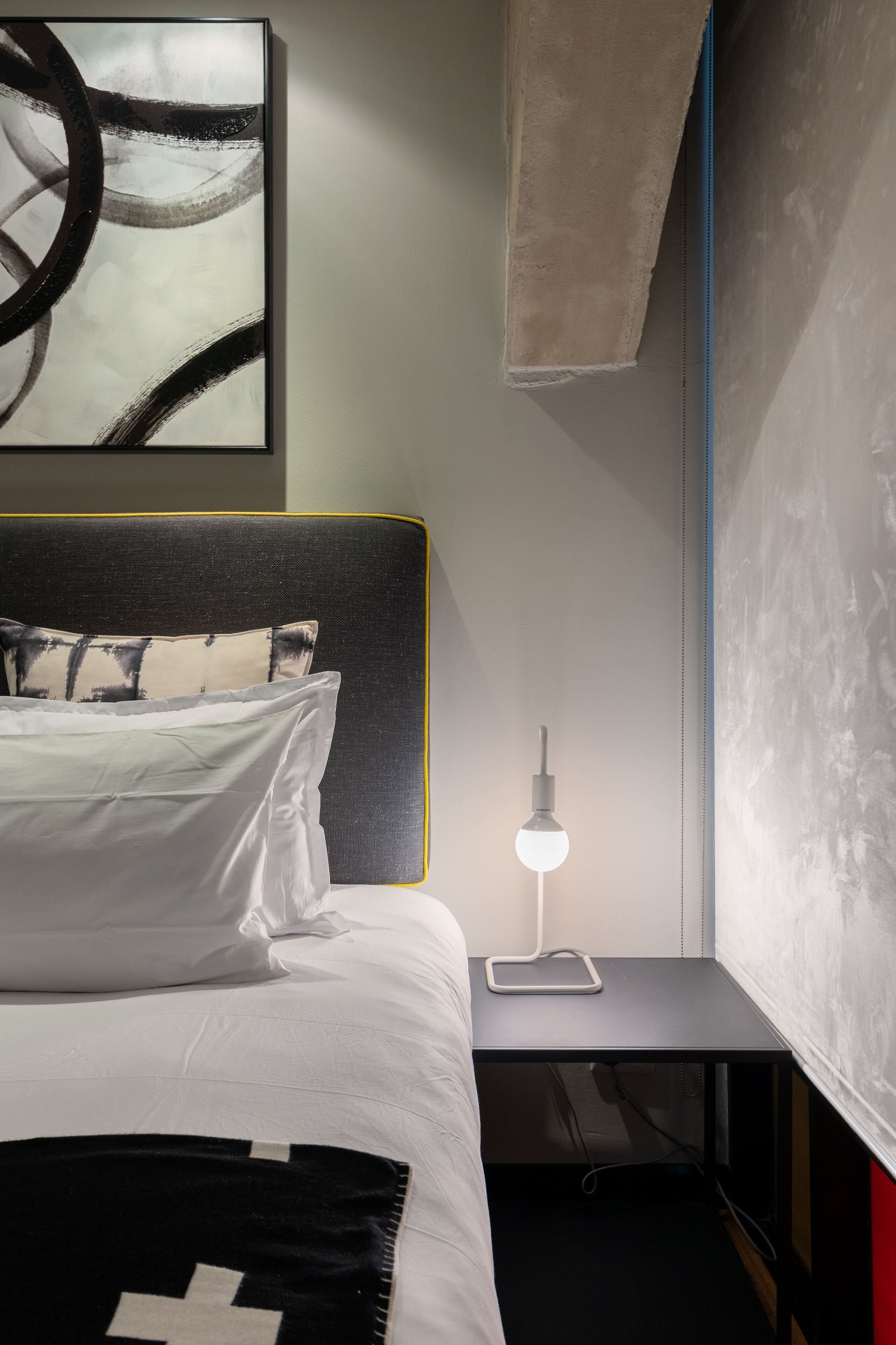 A neatly made bed with white linens and a black and white throw blanket, a bedside table with a lamp, and a wall art piece above the bed.