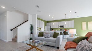 A modern living room with a glass coffee table, a grey sofa, and a red armchair. There is a kitchen area with white cabinets and a green accent wall.