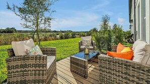 An outdoor patio with wicker furniture, a glass table, and a view of a green field and distant landscape.