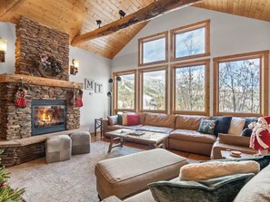 A cozy living room with a stone fireplace, leather sofa, and large windows offering a view of the outdoors. - Amazing Mountain View | Ski In / Ski Out | Hot Tub (Whitefish)