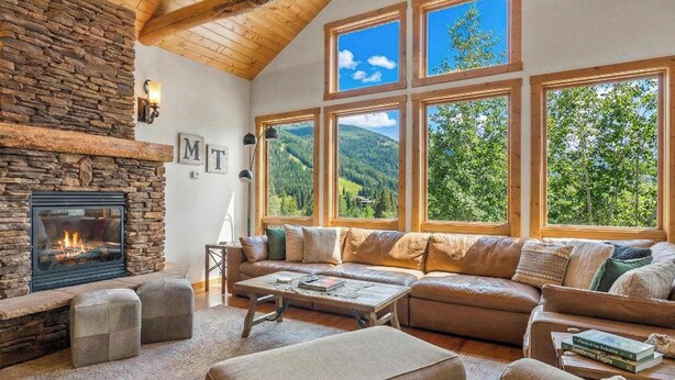 A cozy living room with a stone fireplace, a large sectional sofa, and multiple windows offering views of a forested landscape. - Amazing Mountain View | Ski Access | Hot Tub & Sauna (Whitefish)
