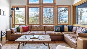 A cozy living room with a large leather sofa, a wooden coffee table, and multiple windows offering a view of a snowy landscape. - Amazing Mountain View | Ski Access | Hot Tub & Sauna (Whitefish)
