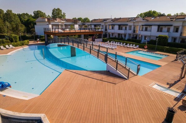 A swimming pool with a wooden deck, lounge chairs, and a building with multiple balconies. - Elegant Flat With Pool and Solarium for six (San Michele al Tagliamento)