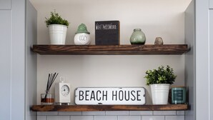 A kitchen shelf with a sign reading "BEACH HOUSE," a white clock, a glass jar with sticks, and potted plants. - Downtown NB Cottage, Backyard, Fire Pit, walkable (New Buffalo)