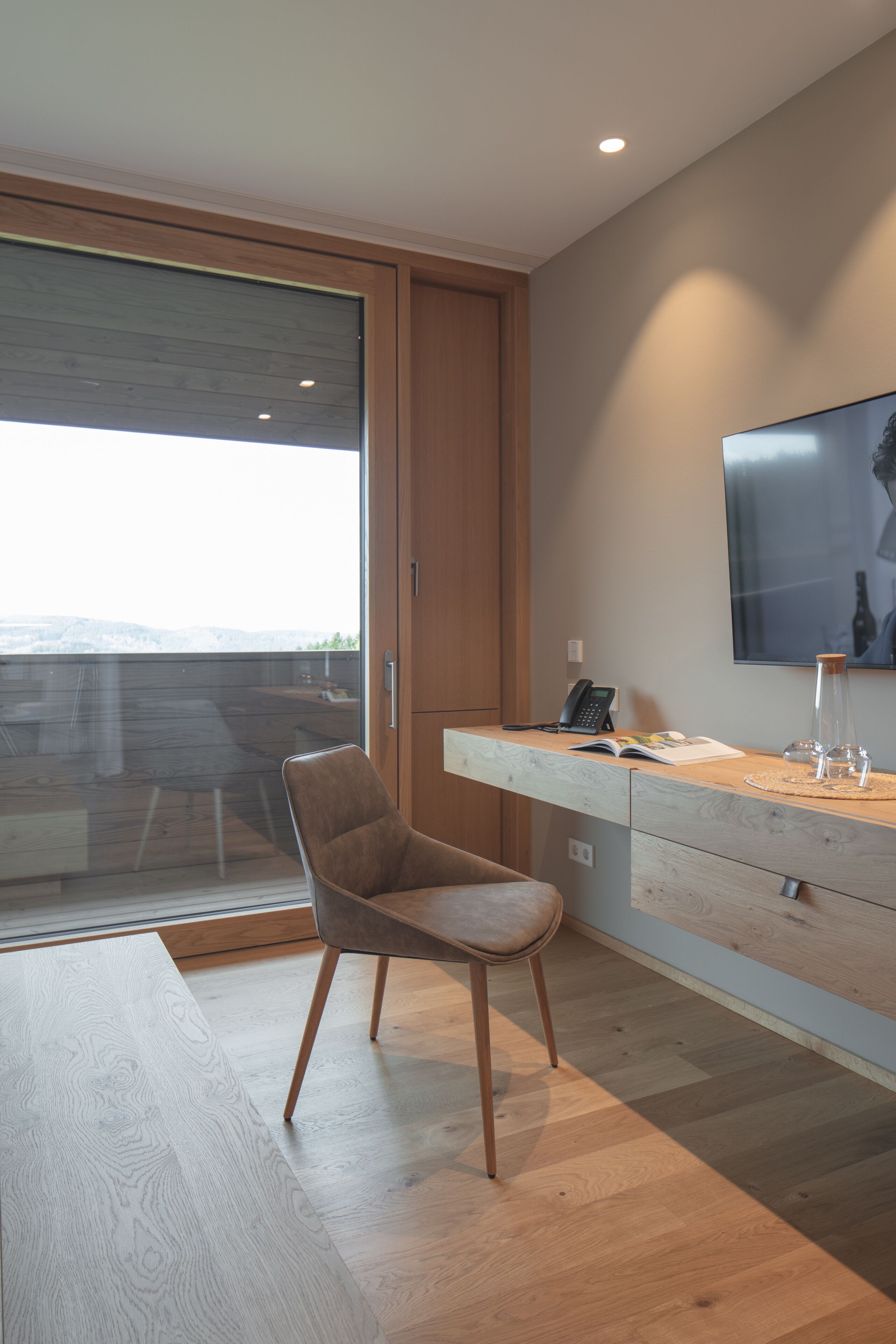 Una habitación moderna con piso de madera, una silla marrón, un escritorio de madera, un televisor montado y un ventanal grande.